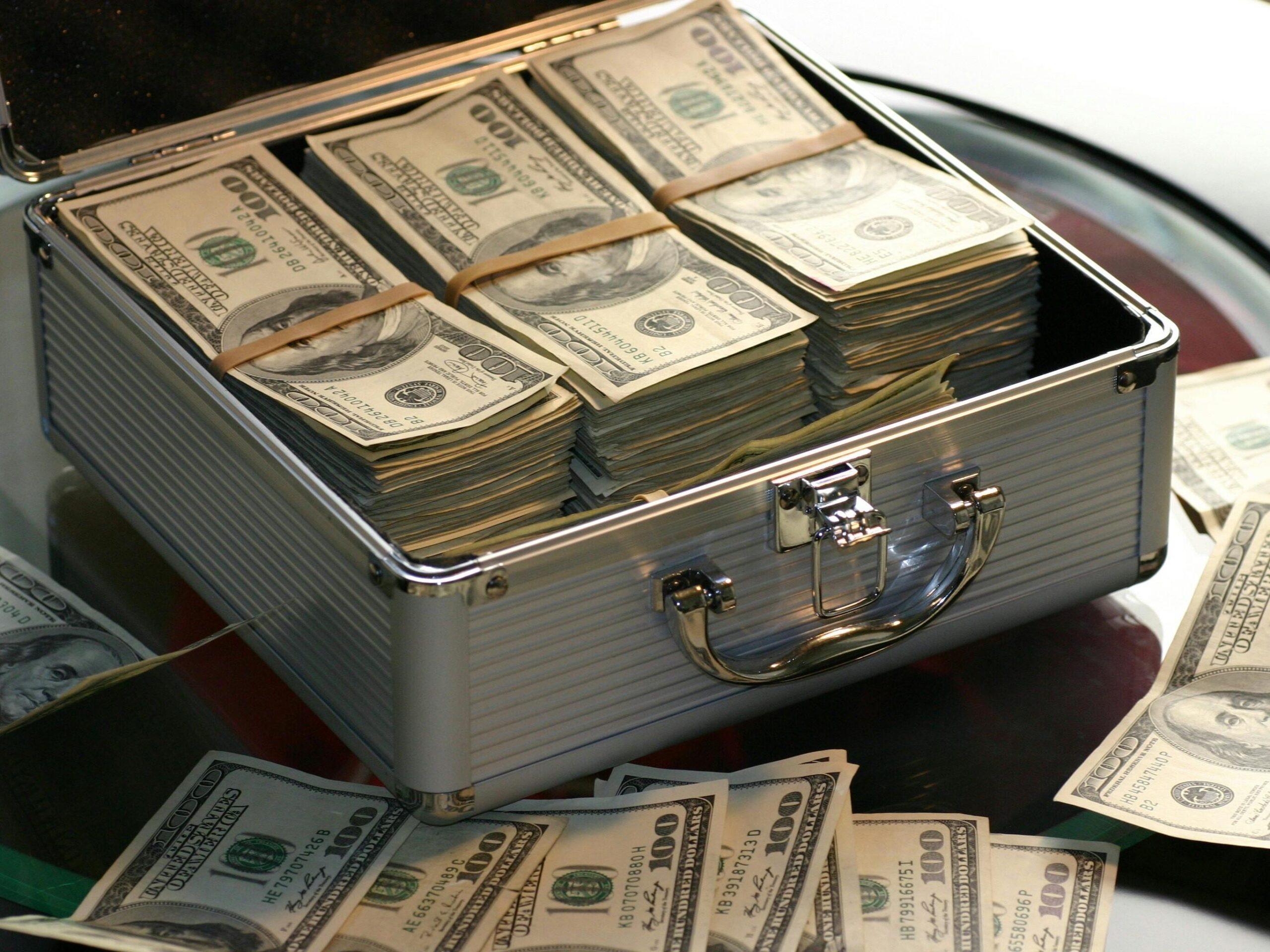 Open briefcase filled with stacks of hundred dollar bills on a glass table, representing wealth.